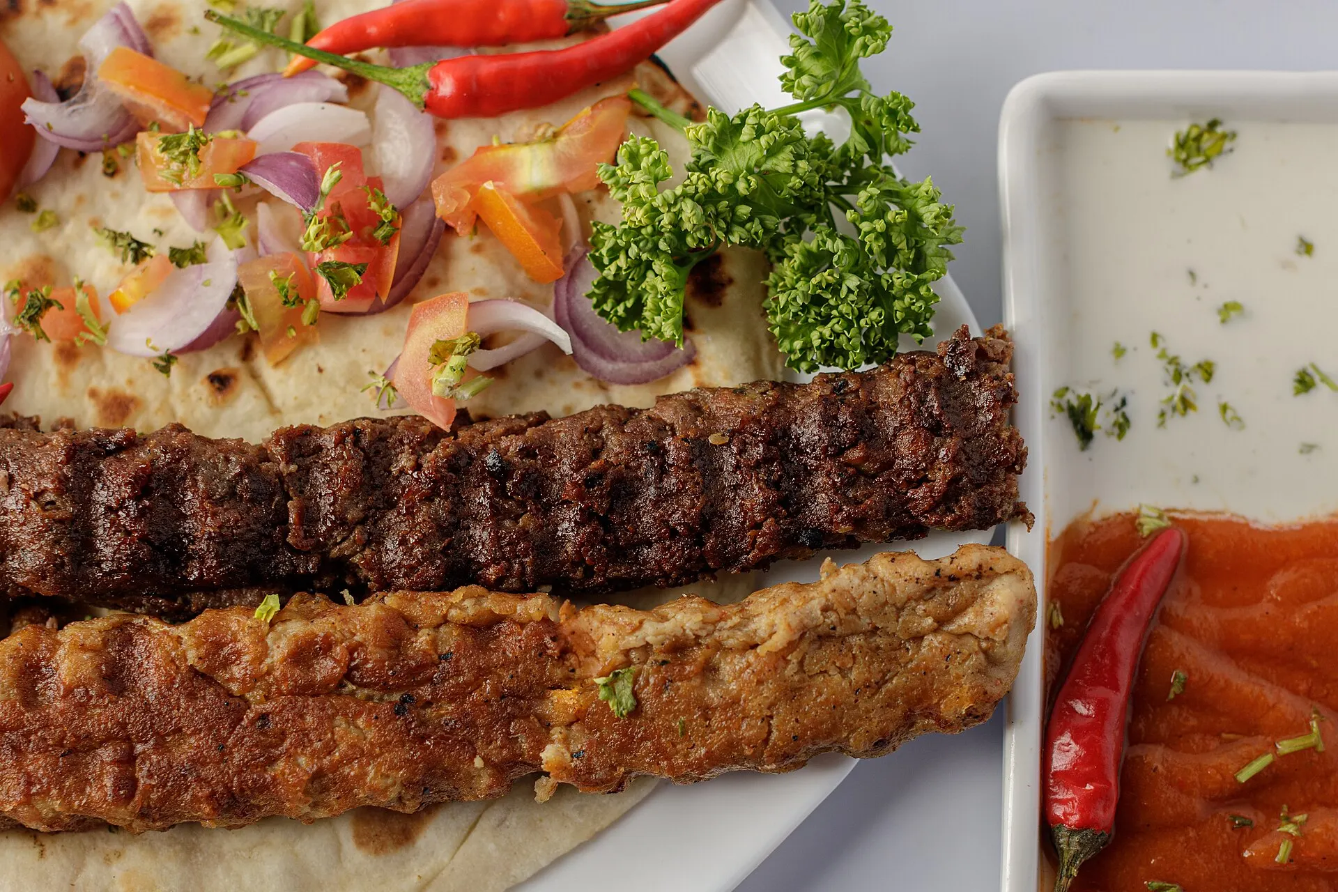 Symbolbild: Gegrillte Kebab-Spieße auf Fladenbrot mit Zwiebeln, Chili und Dips